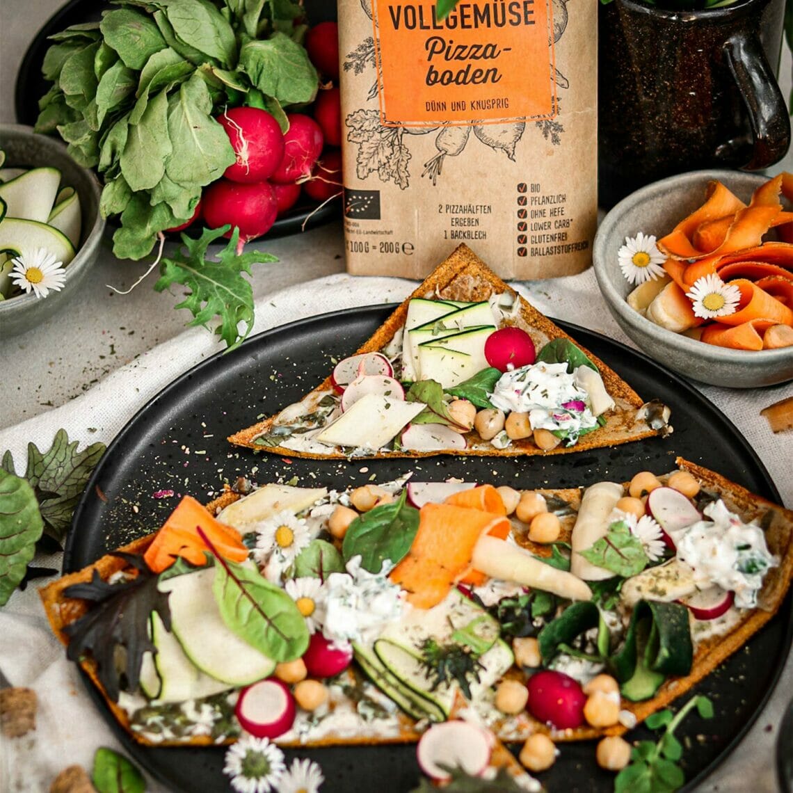 Geschnittener Pizzaboden auf schwarzem Teller belegt mit Kichererbsen, Spargel, Zucchini, Radieschen, Karotten, Salat und Gänseblümchen